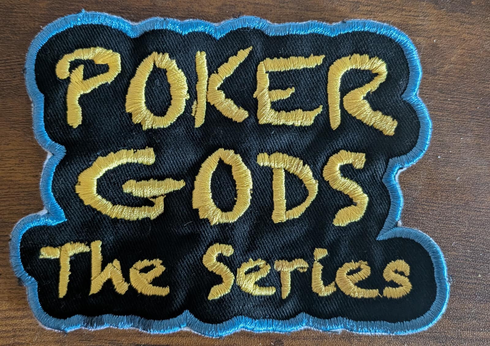 Embroidered Poker Gods: The Series patch with gold lettering on black twill and a blue stitched border
